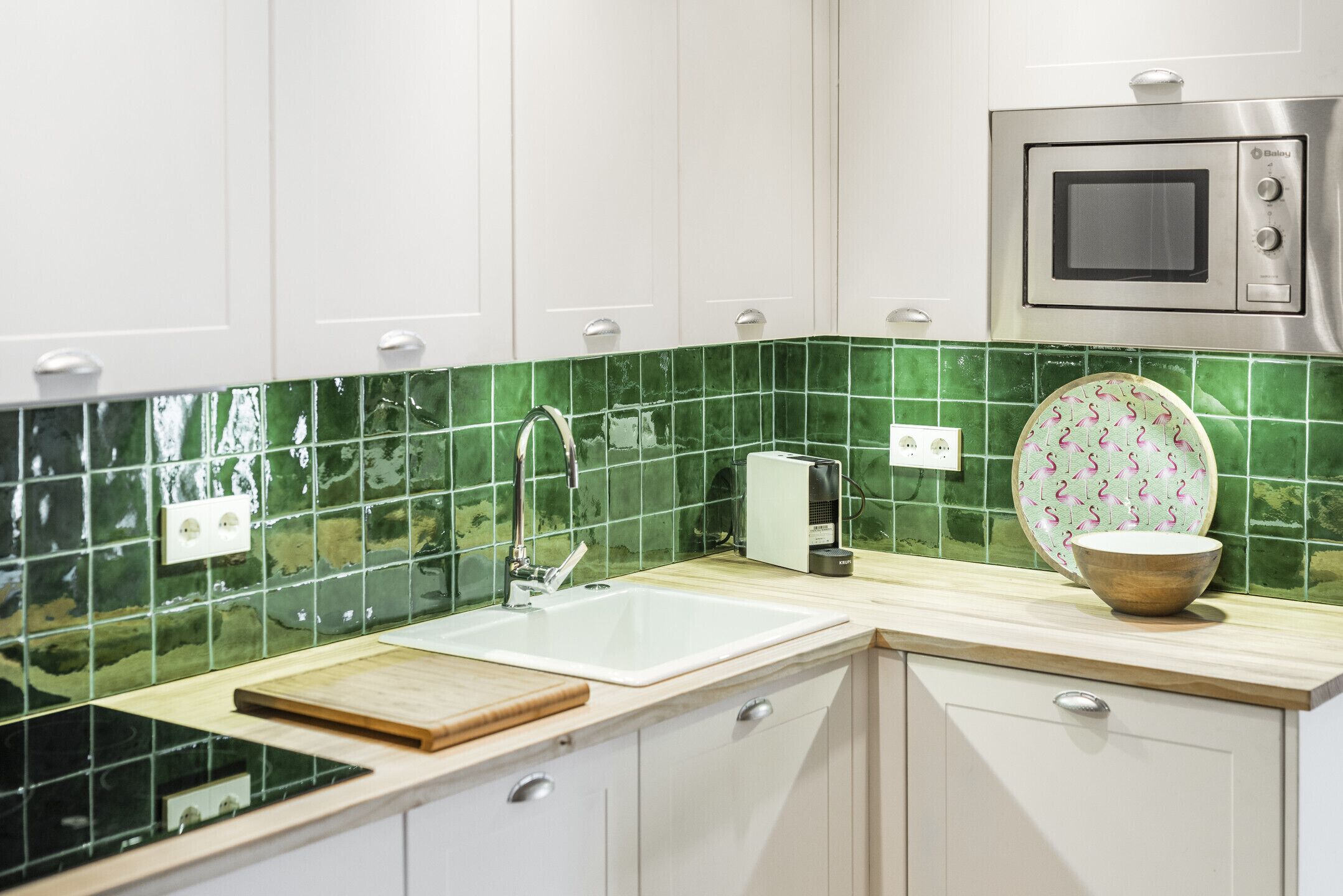 A kitchen with white cabinets, a green tiled backsplash, a sink, a microwave, and a wooden cutting board.