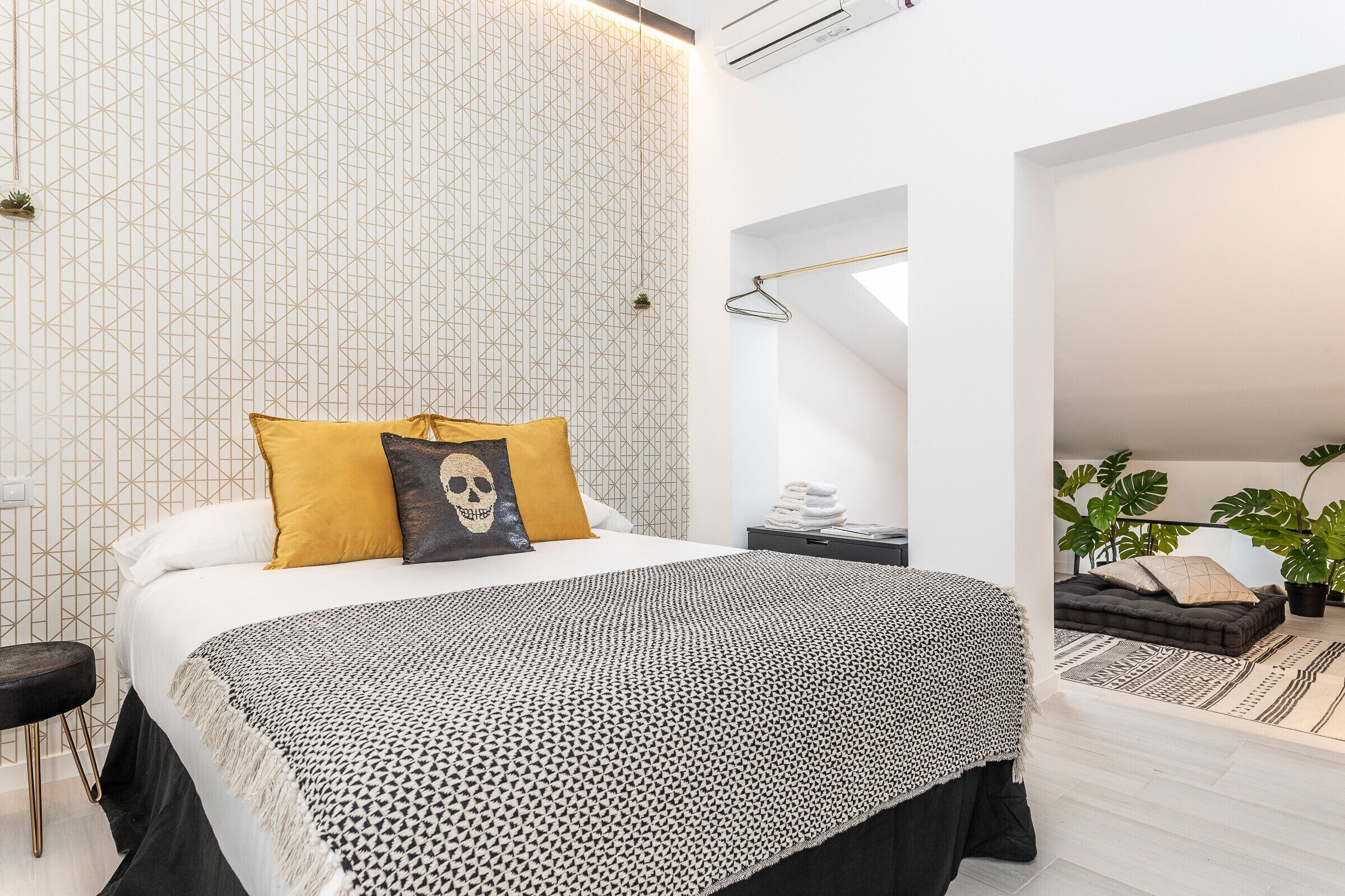 Un dormitorio moderno con papel tapiz estampado con motivos geométricos, una cama con un cojín con temática de calavera y una manta de diseño blanco y negro.