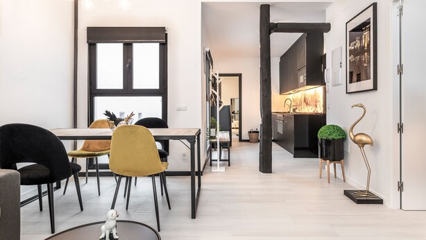A modern dining area with a wooden table, black and yellow chairs, and a small white dog on the floor. - Waou Malasaña es MIA - Corta Duración (Madrid)
