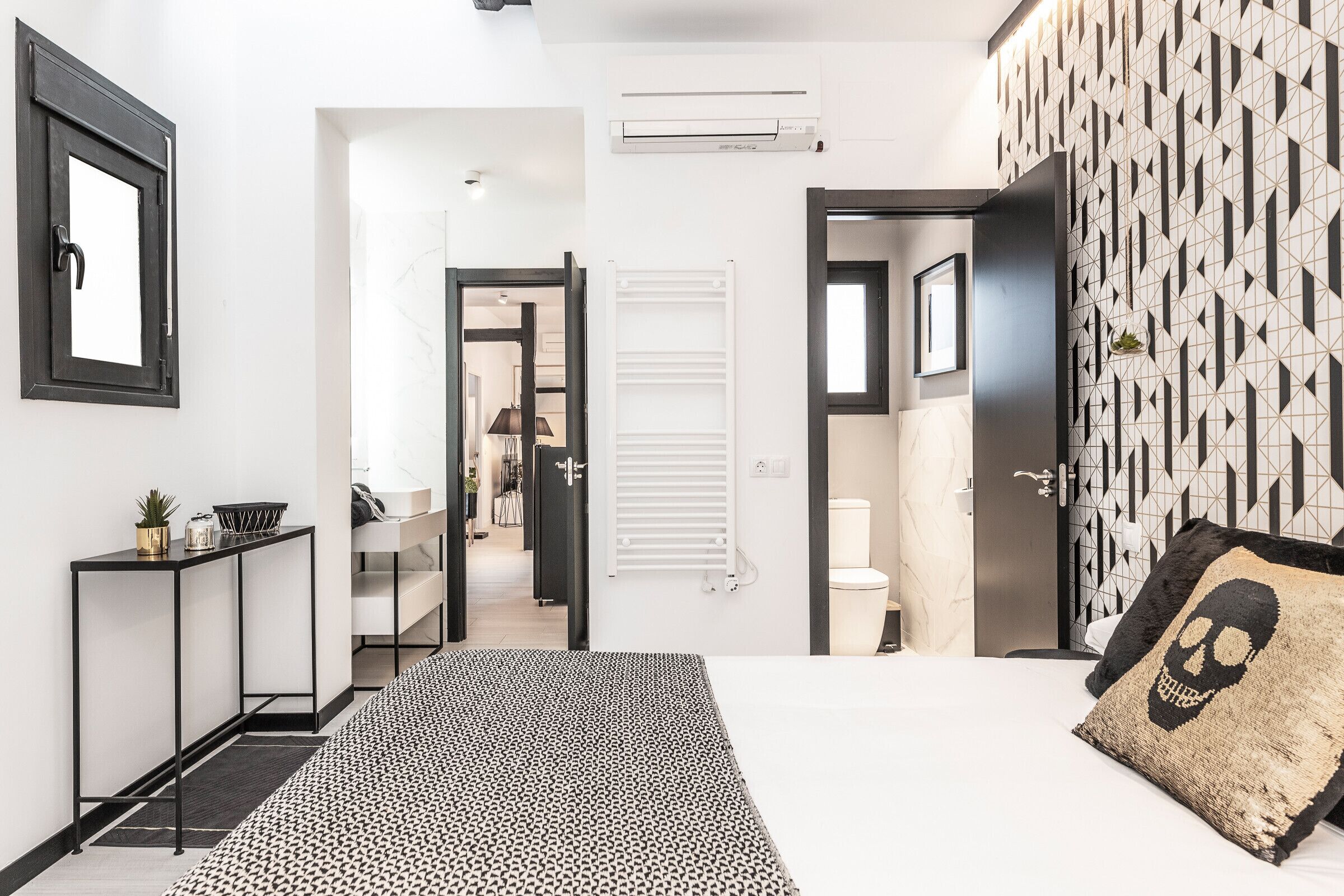 A modern bedroom with a bed, a console table, a patterned rug, a geometric wall design, and a door leading to another room.