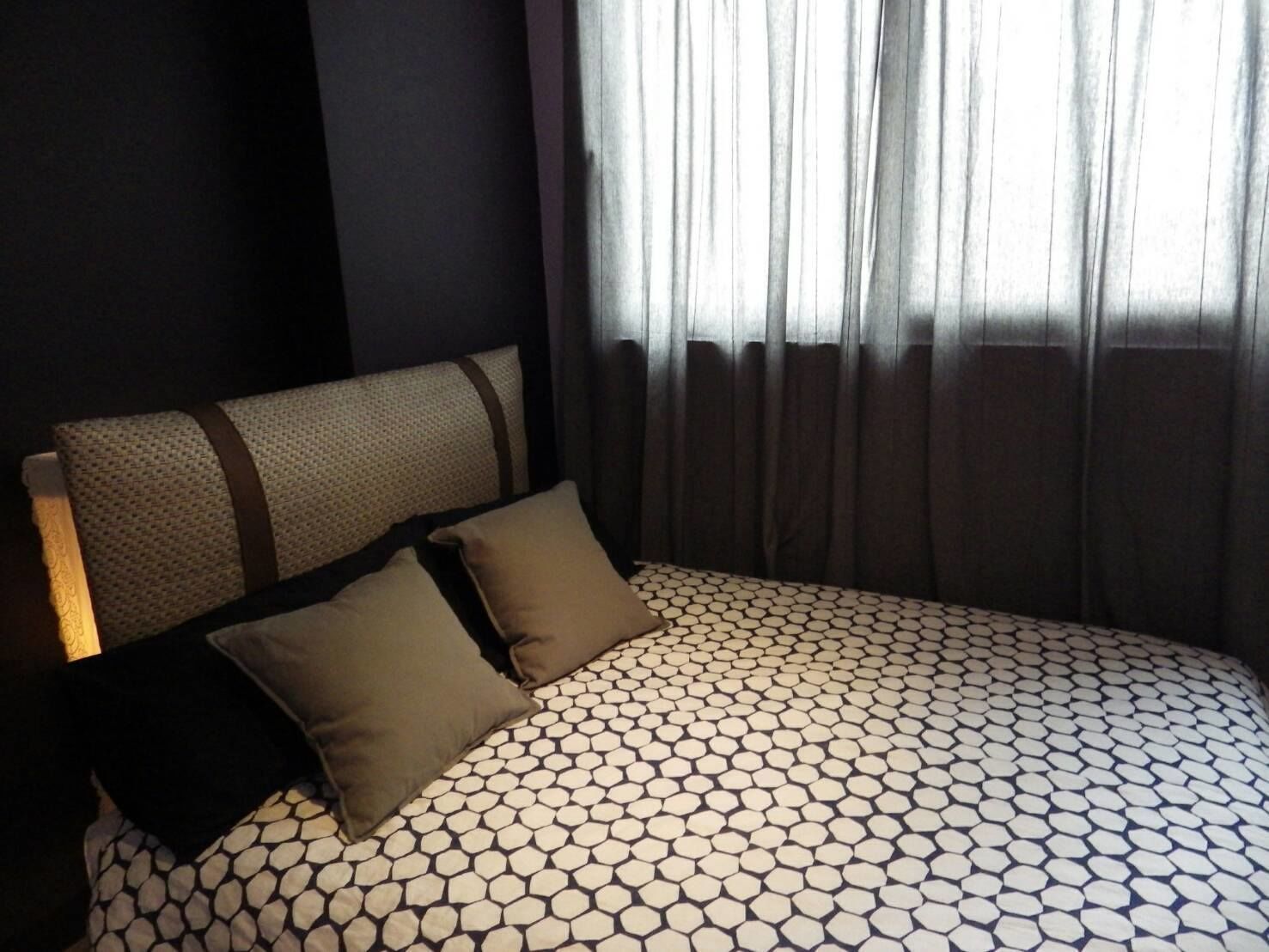 A hotel room with a bed featuring a geometric patterned bedspread, a headboard with a textured fabric, and a window with sheer curtains.