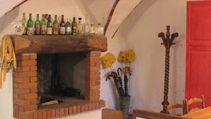 Ein Kamin aus Ziegelsteinen mit einem Regal darüber, auf dem verschiedene Flaschen stehen. Rechts vom Kamin befindet sich ein mit gelben Blumen gefüllter Strauß. Ein Holzstuhl ist im Vordergrund zu sehen.
