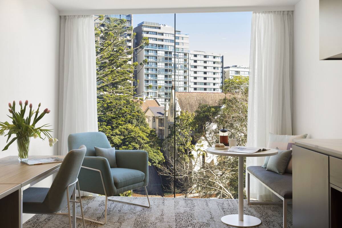 Habitación moderna con vistas a edificios altos y árboles. Cuenta con un escritorio, una silla, un sofá y una pequeña mesa con una taza y un libro.
