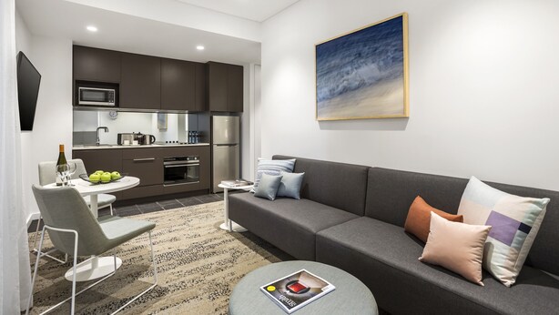 A modern living room with a grey sofa, a round coffee table, and a dining area with a white table and chairs. - Quest North Sydney (North Sydney)