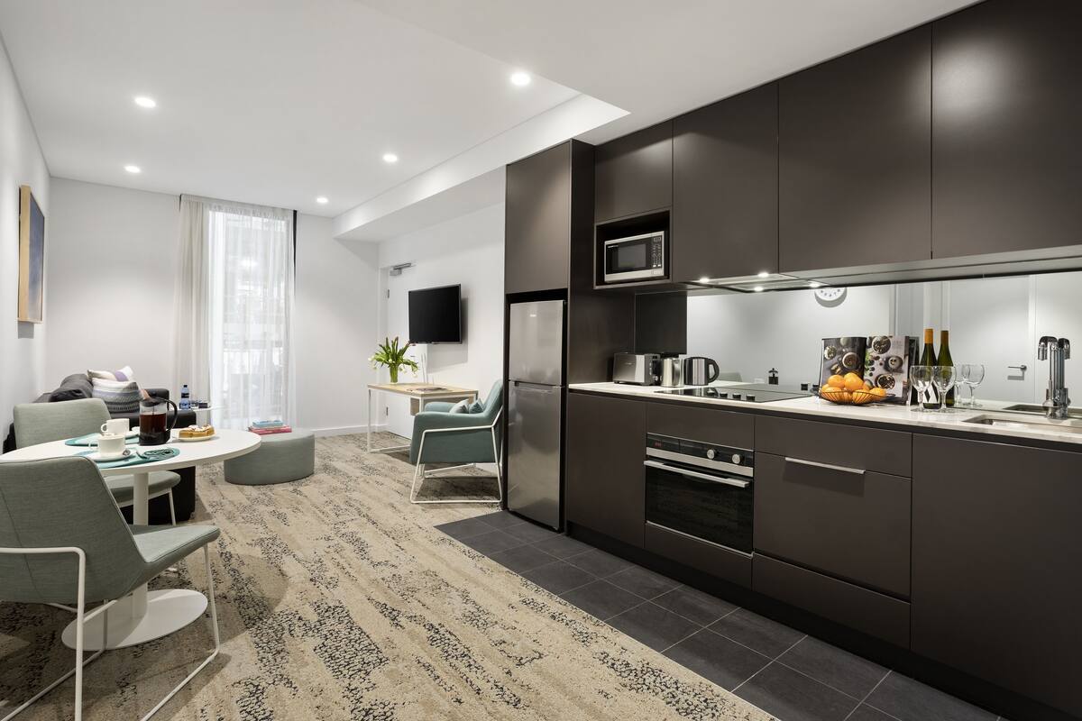 Una cocina moderna con armarios oscuros, microondas y horno empotrado. Hay una zona de comedor con mesa y sillas, y un amplio ventanal con cortinas translúcidas.
