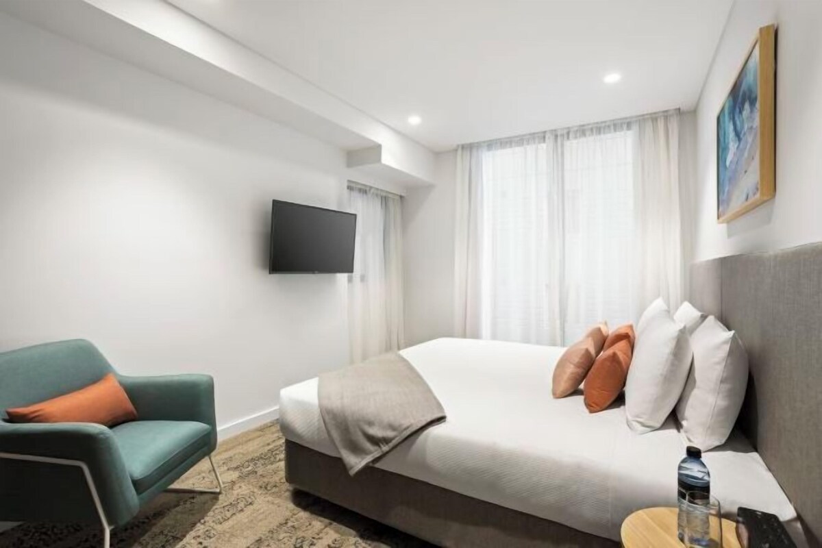 A hotel room with a bed, a chair, a TV mounted on the wall, and a painting on the wall.