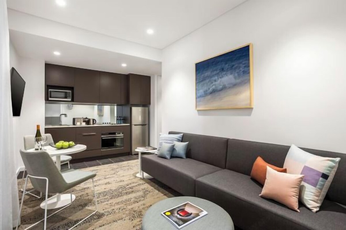 Una sala moderna con un sofá gris, una mesita redonda pequeña y un comedor con una mesa y sillas blancas. Se ve una cocina al fondo con armarios oscuros y electrodomésticos modernos.