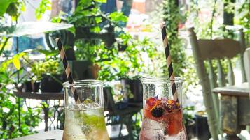 Two glass bottles with straws on a wooden table, one with a brown liquid and the other with a red liquid, both with ice and garnishes, in a sunny outdoor setting with greenery in the background.