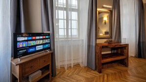 Een kamer met een houten tv-meubel, een groot raam met dunne gordijnen, een spiegel en een houten tafeltje.