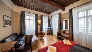 Een hotelkamer met een houten plafond, een bank, een bed met een rode deken, een houten tafel, een stoel, een televisie en een raam met gordijnen.