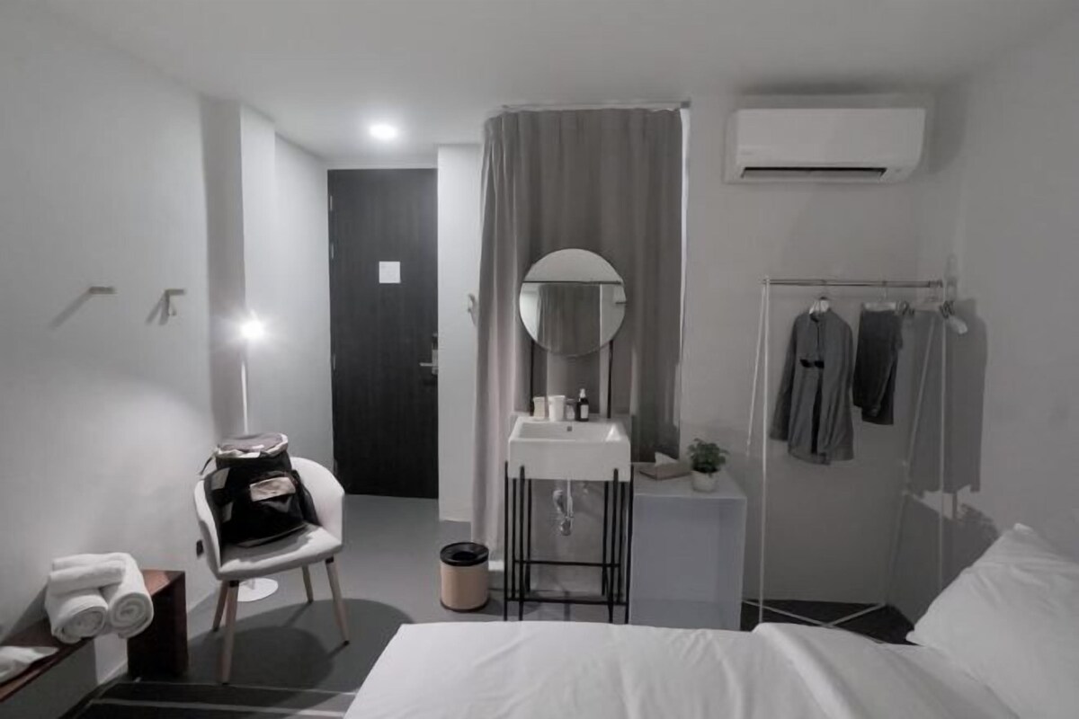 A modern hotel room with a bed, a small desk, a chair, a vanity with a mirror, a shower area, and a hanging clothes rack.