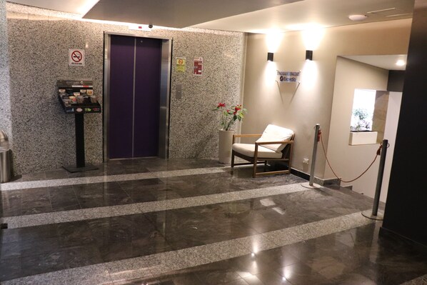 A hotel lobby with a marble floor, a vending machine, a purple elevator, a waiting area with a chair, and a wall-mounted sign.