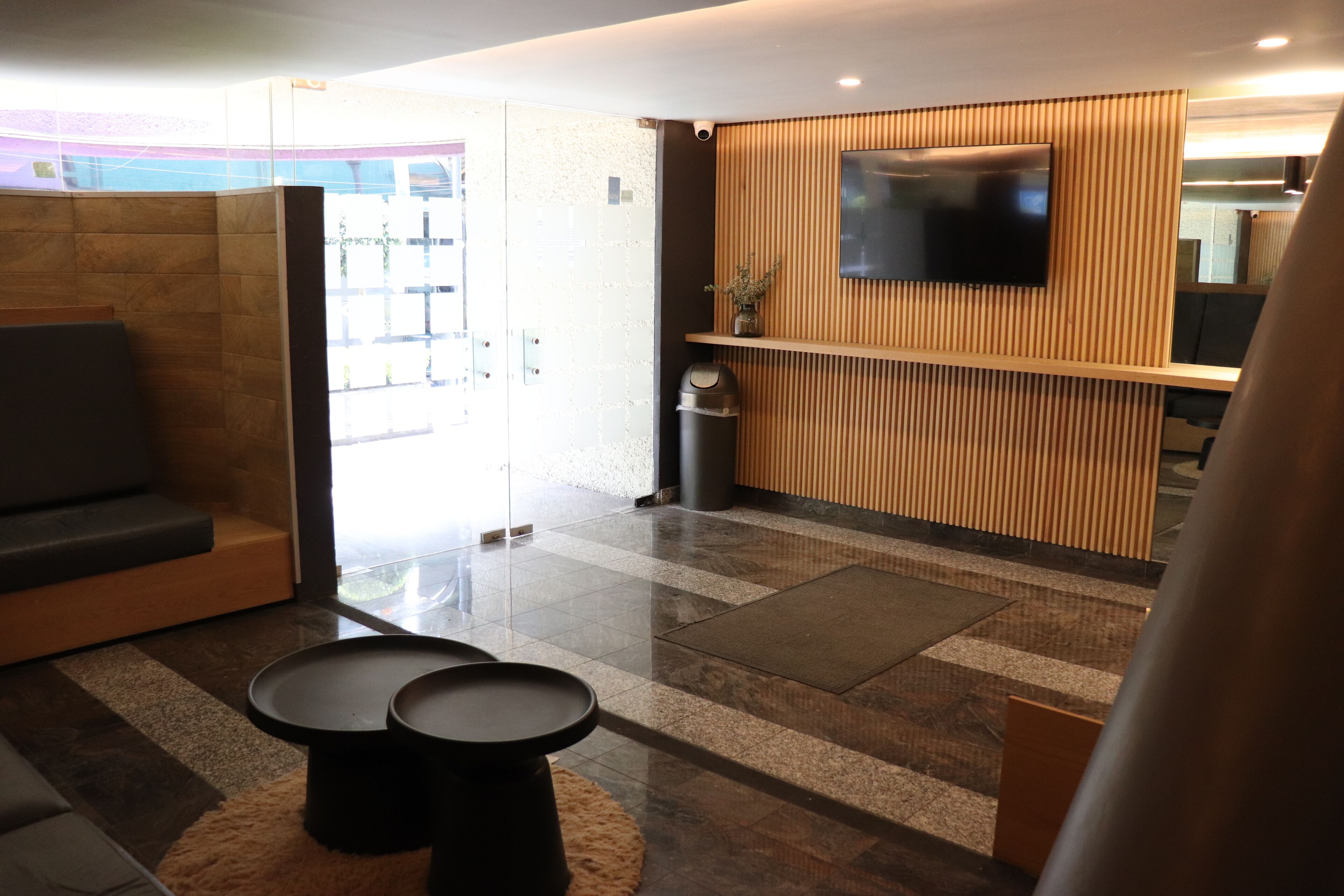 Un hall d’entrée avec un canapé, deux tables rondes, un comptoir d’accueil en bois et un téléviseur fixé au mur.