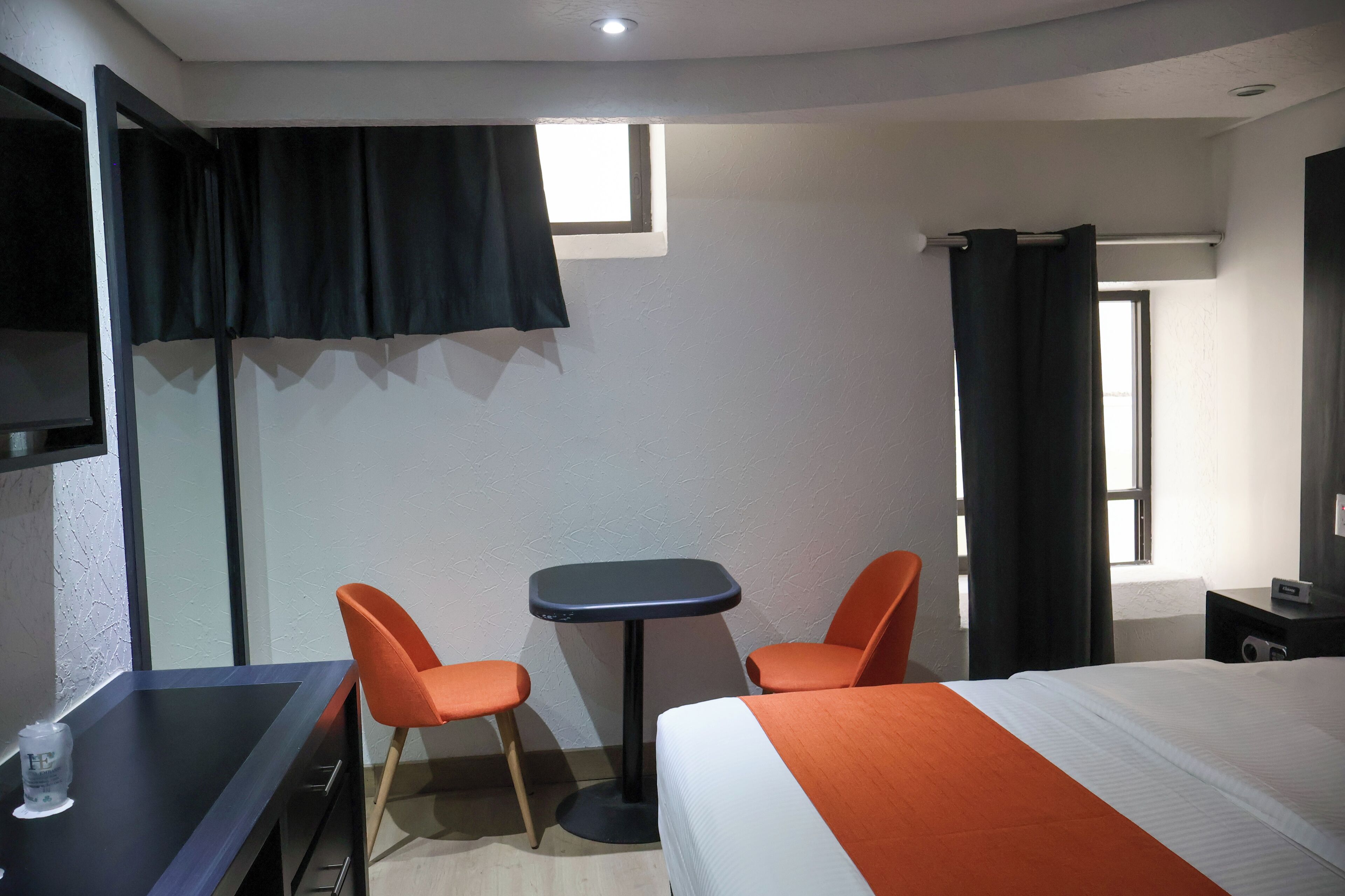 A hotel room with a bed, two orange chairs, a small table, a TV, and a window with curtains.