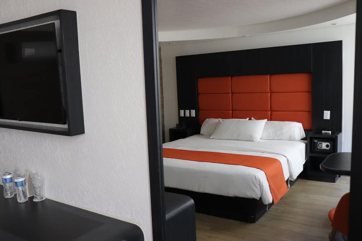 Habitación de hotel con una cama grande, un televisor montado en la pared, una mesita de noche con lámpara y una pequeña mesa con agua embotellada.