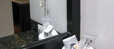 A hotel bathroom with a hairdryer mounted on the wall, a towel dispenser, and a trash bin with a towel.