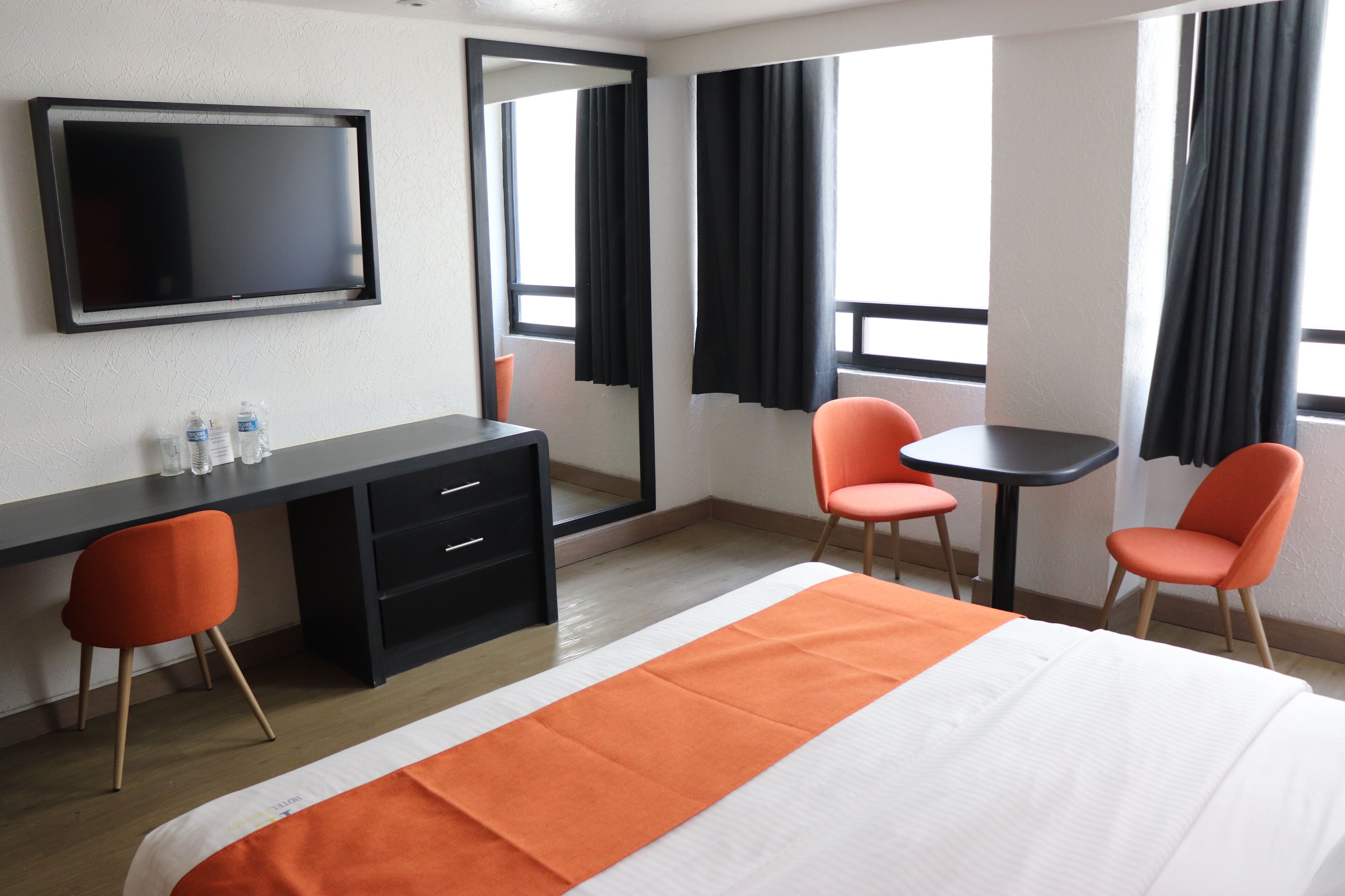 Une chambre d’hôtel avec un lit, un bureau, deux chaises, une télévision et une fenêtre avec des rideaux.