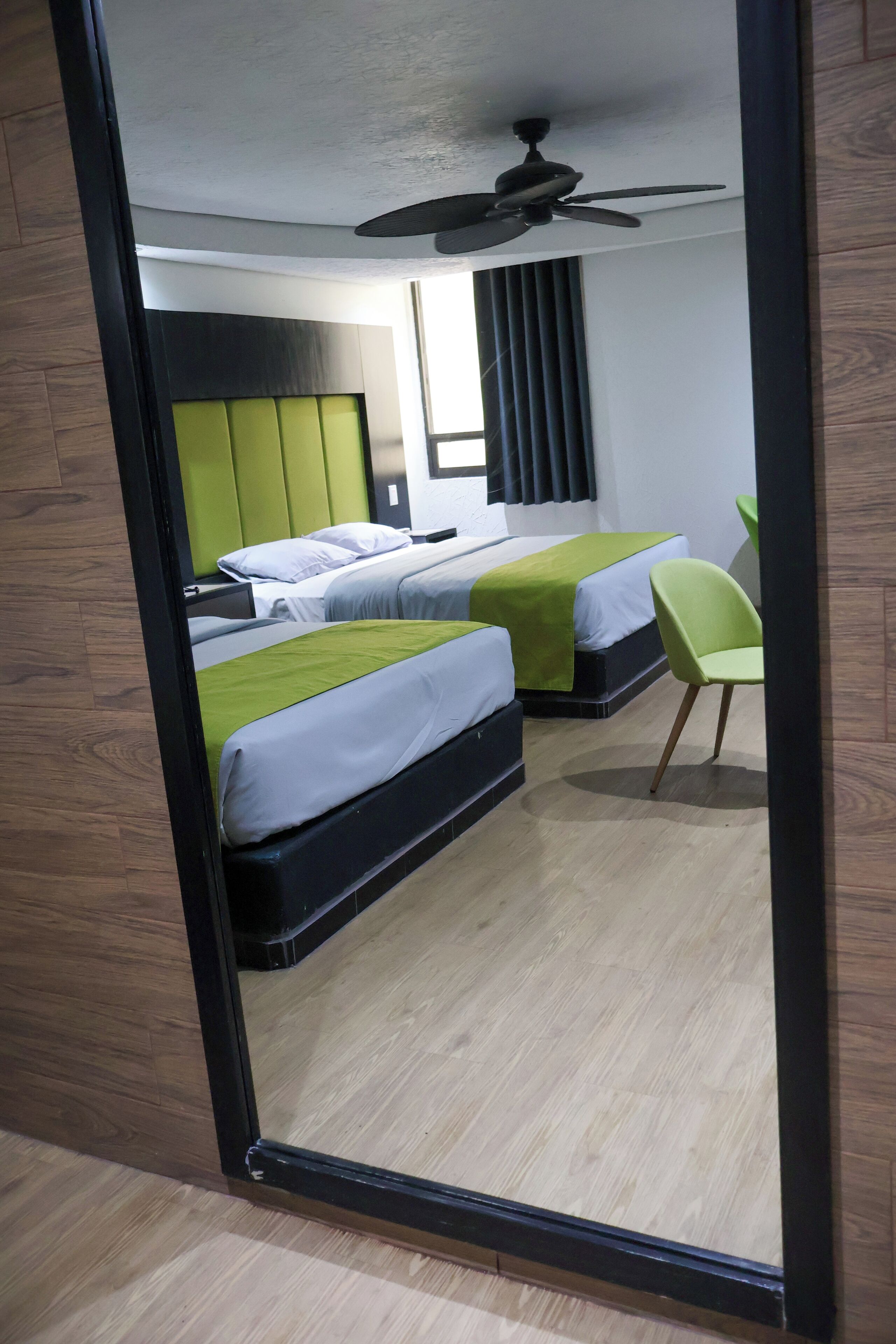A hotel room with two beds, a ceiling fan, a green chair, and a window with curtains.