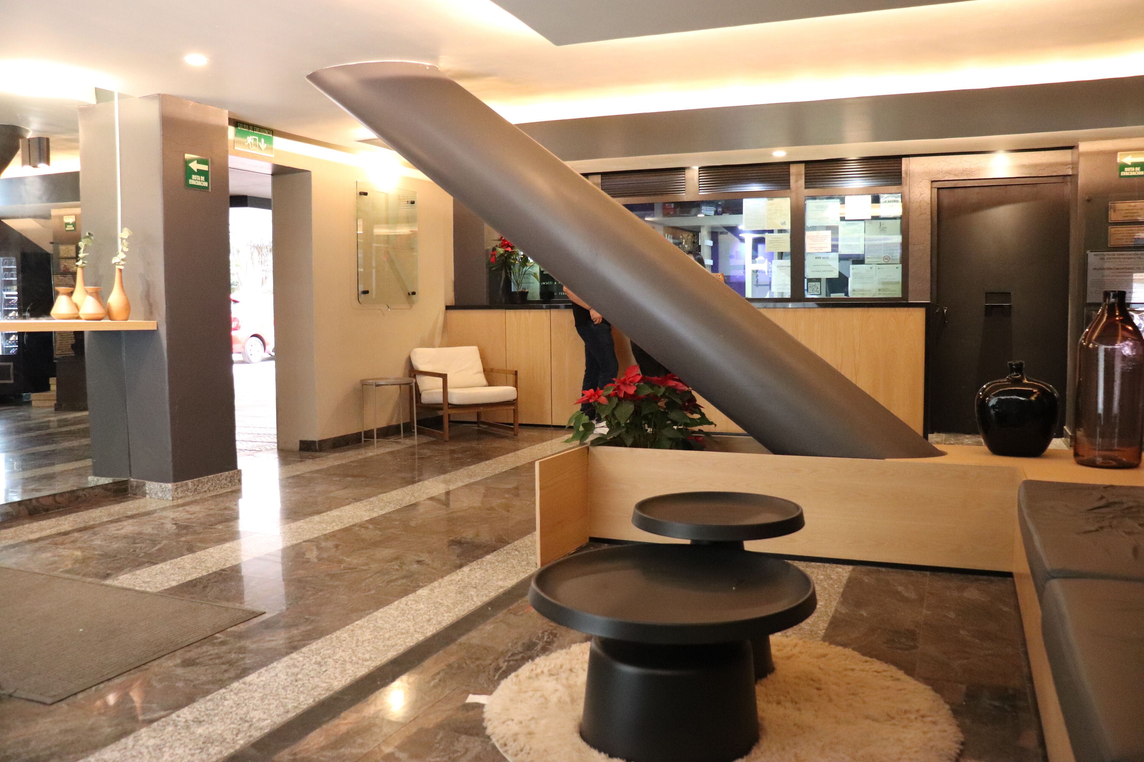 Un vestíbulo con diseño moderno, que presenta una gran columna cilíndrica, un mostrador de recepción y una zona de estar con una mesita y una silla.