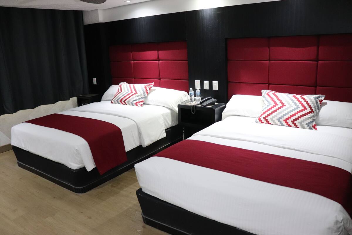 Habitación de hotel con dos camas, cada una con ropa de cama blanca y detalles rojos, cabecero negro y mesita de noche con teléfono y botellas de agua.