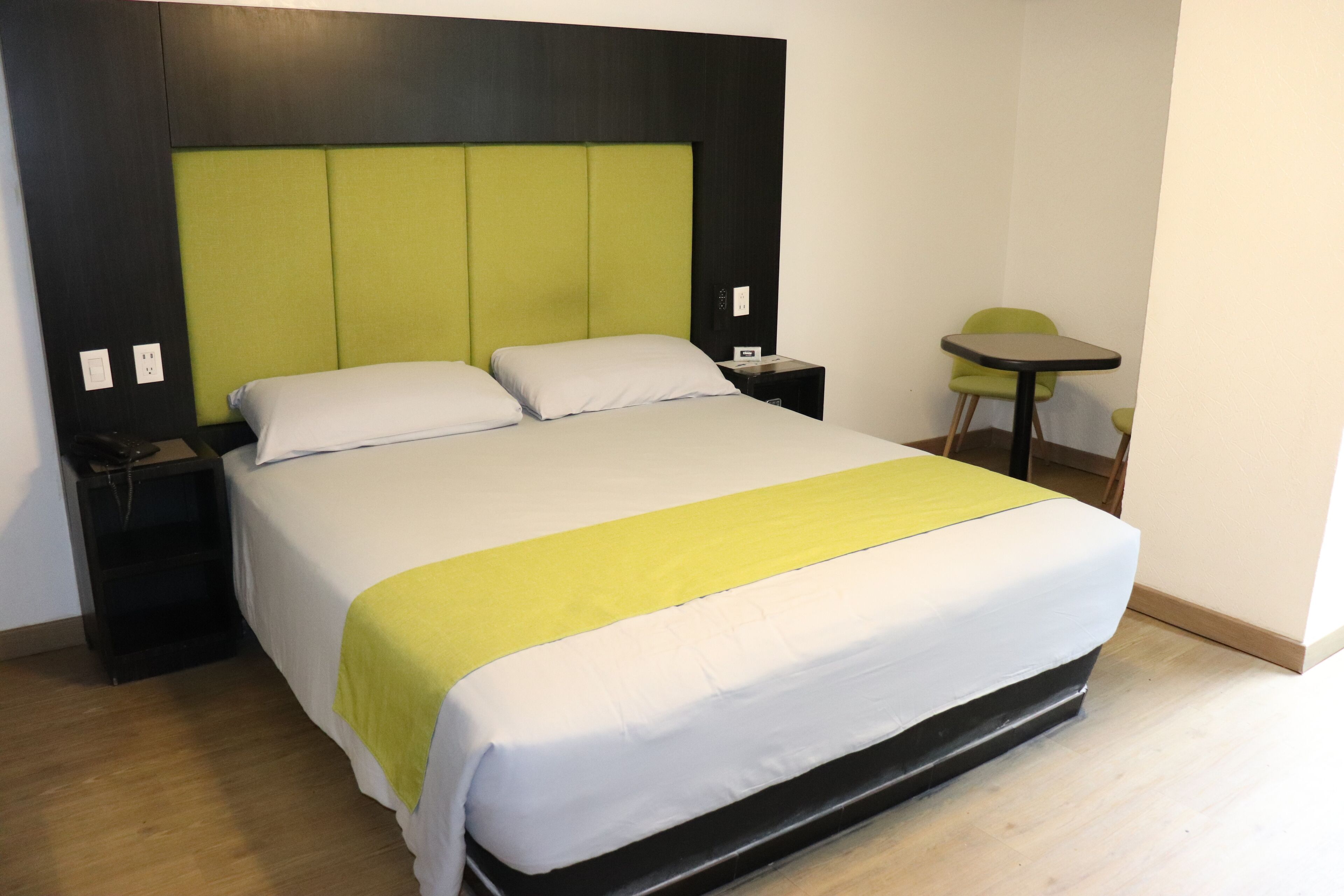 A hotel room with a large bed, a green headboard, a small table, and a chair.