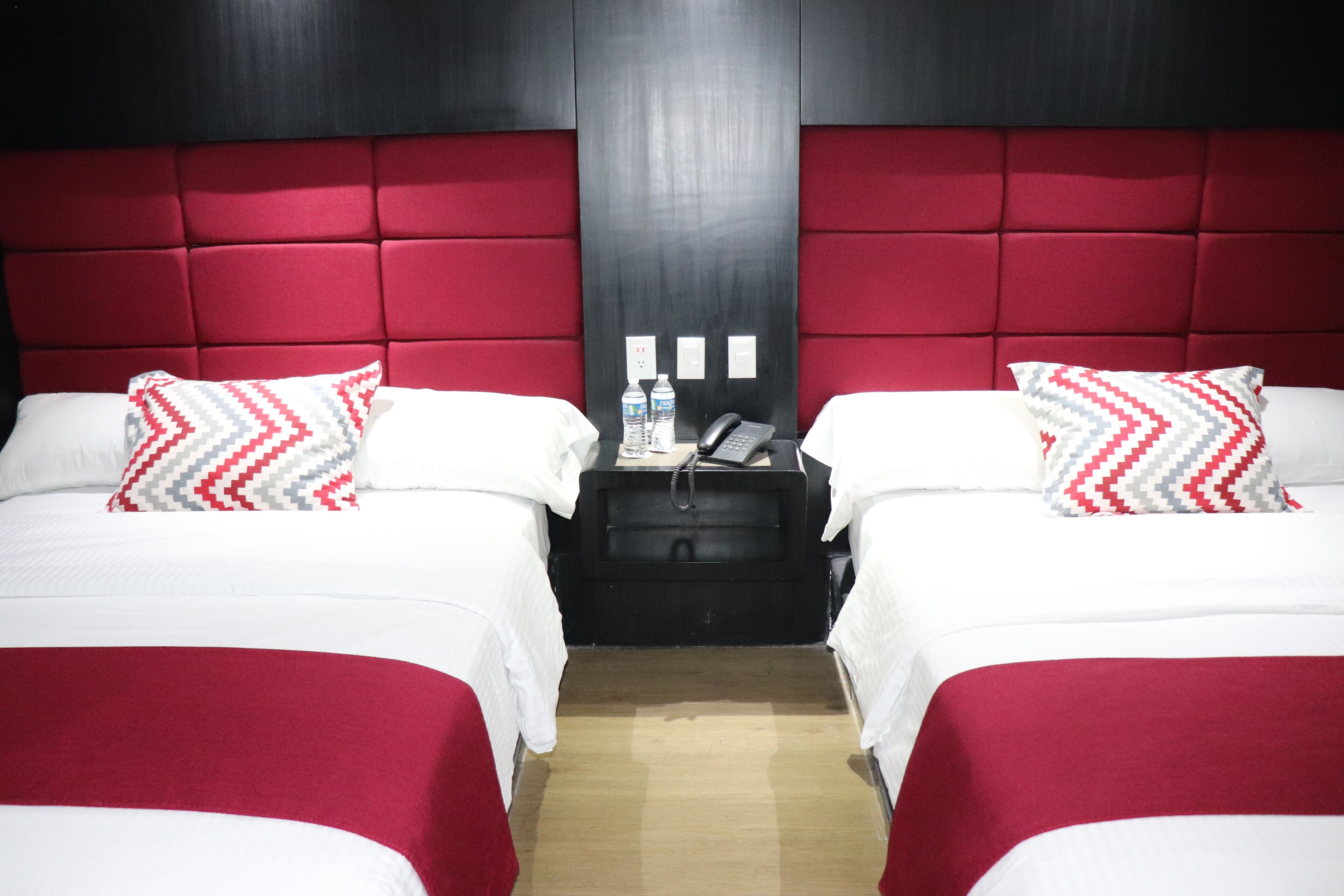 Dos camas individuales con ropa de cama roja y blanca, una mesita de noche negra con un teléfono y una botella de agua, y un cabecero rojo.
