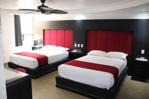 Habitación de hotel con dos camas, cada una con ropa blanca y detalles rojos, un ventilador de techo y una mesita de noche con teléfono.