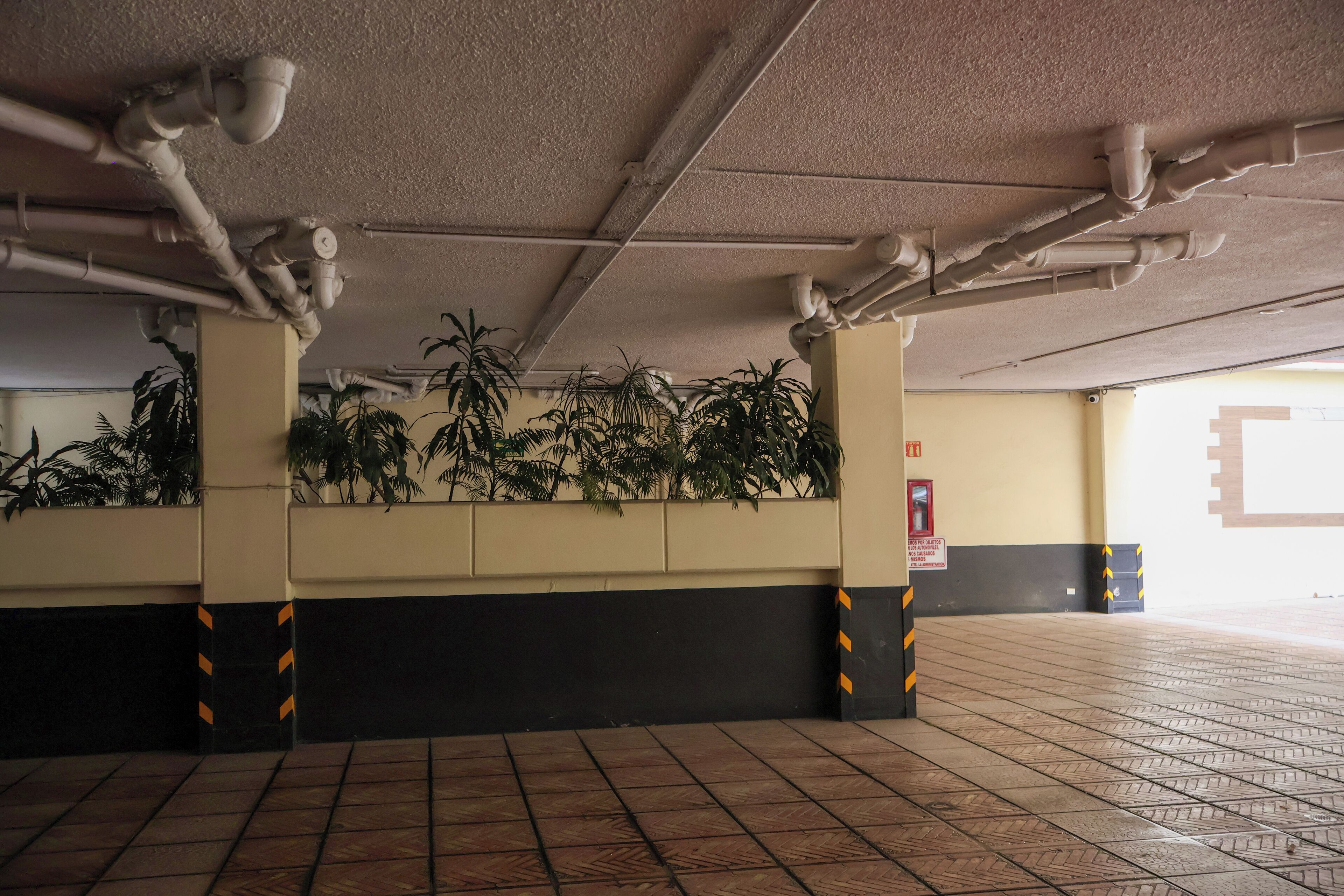 Un estacionamiento interior con suelo de baldosas, tuberías en el techo y plantas en macetas a lo largo de las paredes.