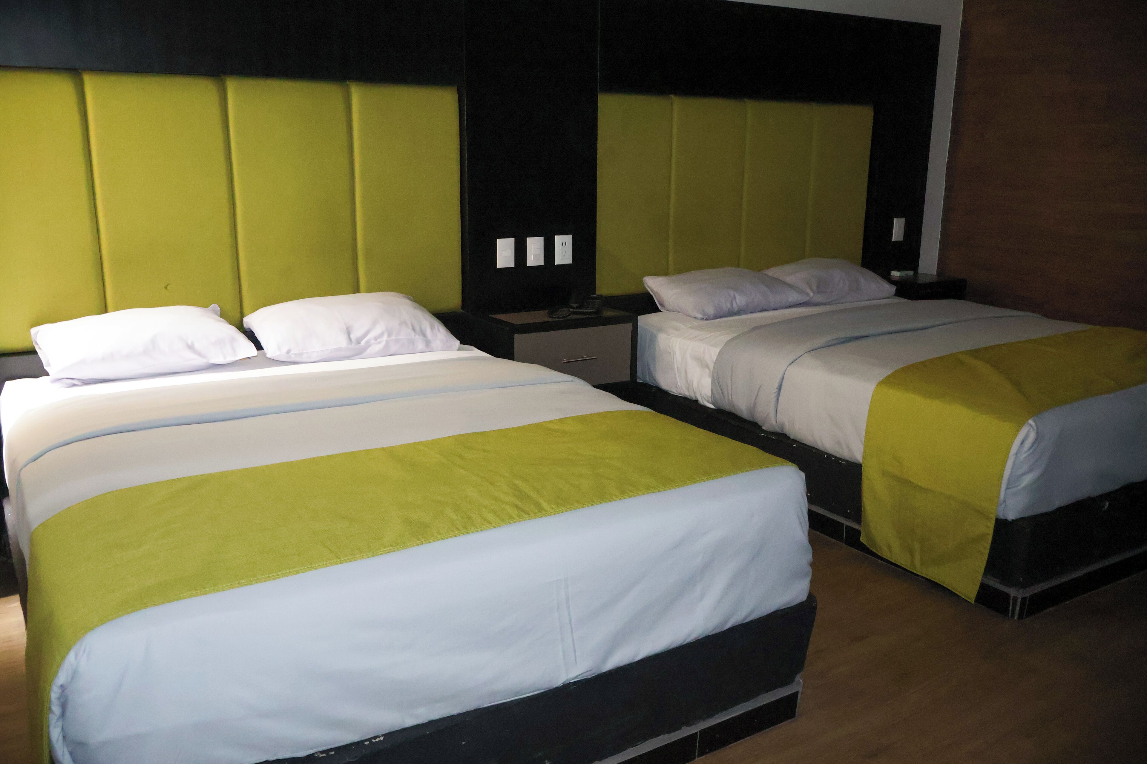 A hotel room with two beds, each with a green and white bedspread, a wooden floor, and a modern headboard design.