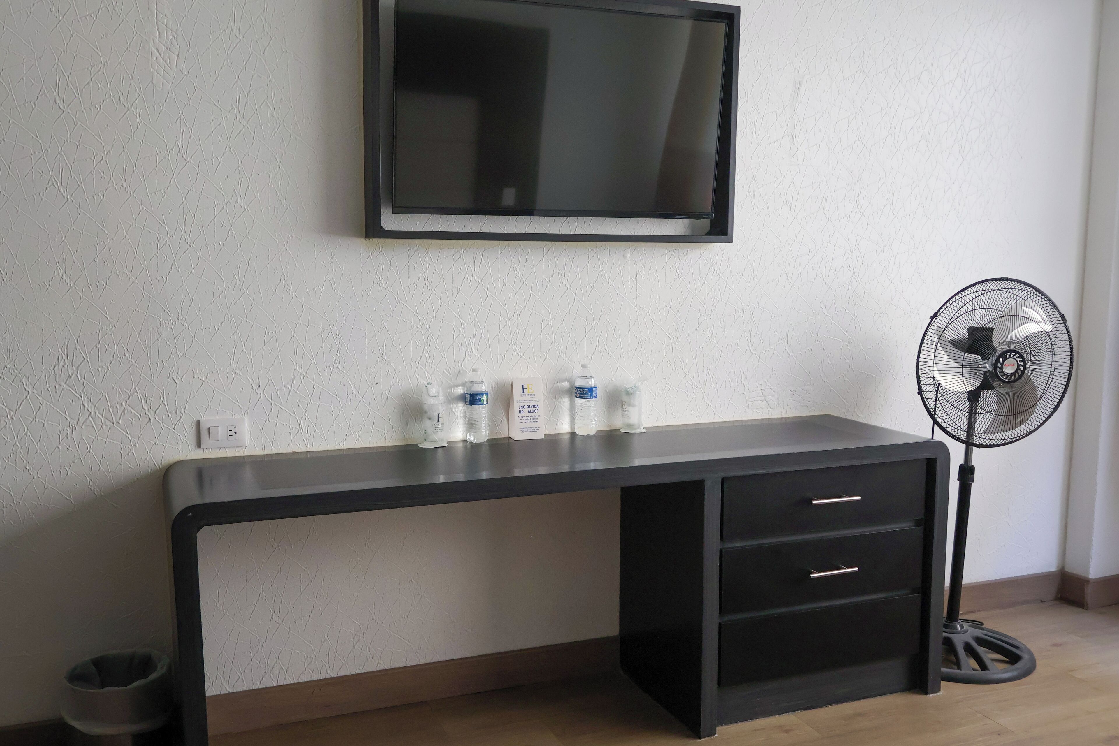 Une pièce avec un bureau noir, un téléviseur fixé au mur et un ventilateur sur pied.