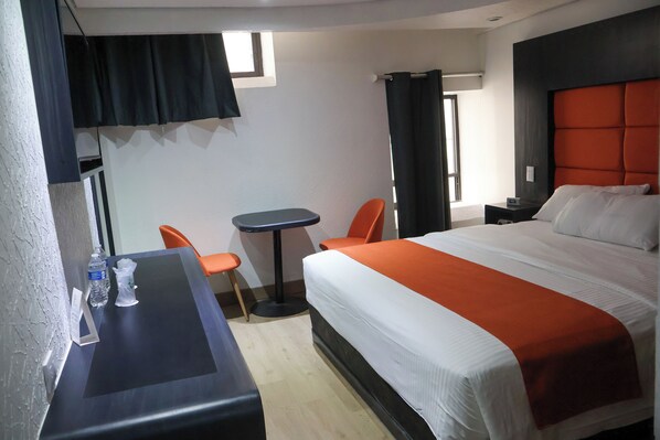 Habitación de hotel con una cama grande, un escritorio con silla, una mesita y una ventana con cortinas.