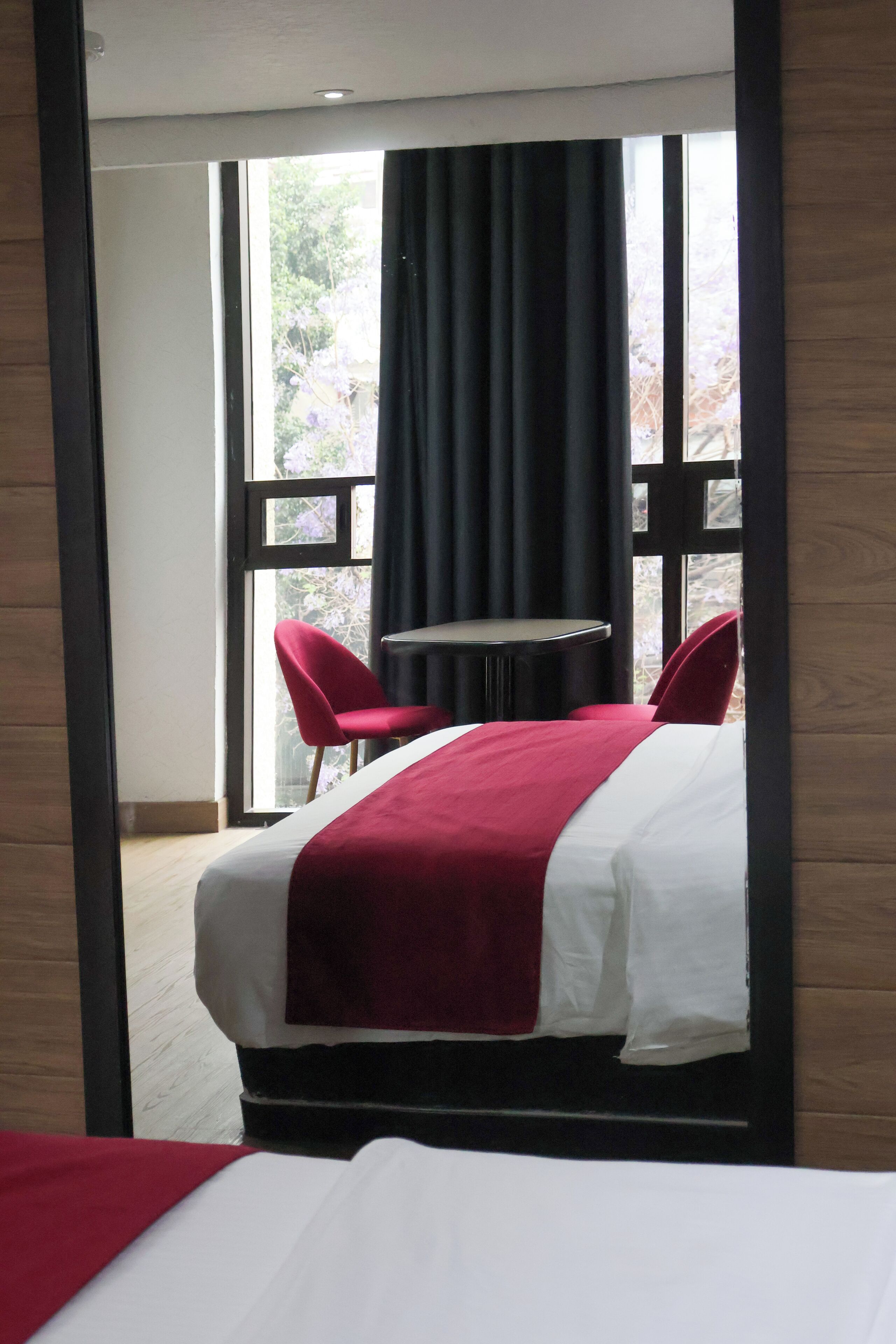 A hotel room with a bed, a small table, two red chairs, and a large window with curtains.