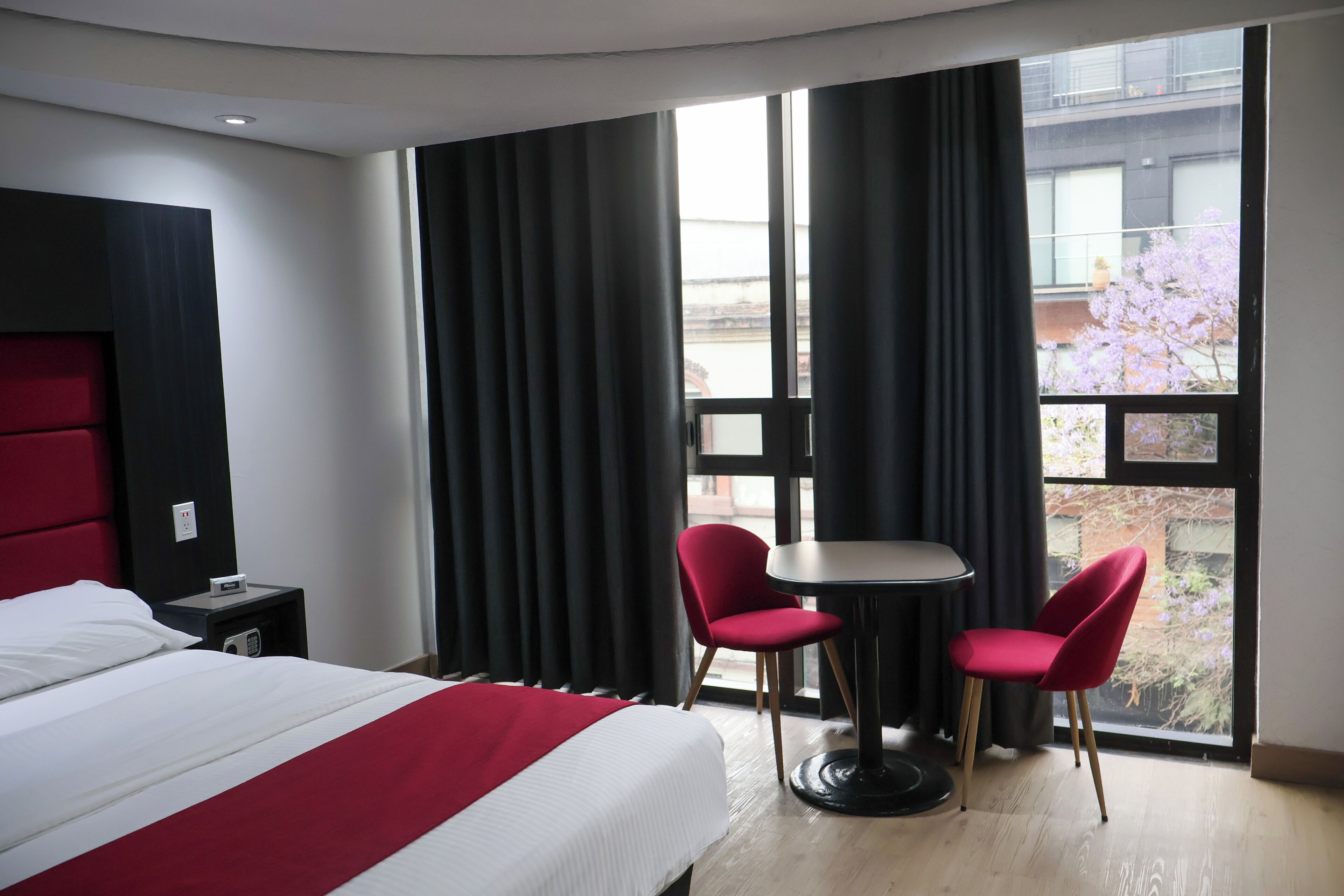 Une chambre d’hôtel avec un lit, une table, deux chaises, une fenêtre avec des rideaux et une vue sur un immeuble et des cerisiers en fleurs à l’extérieur.