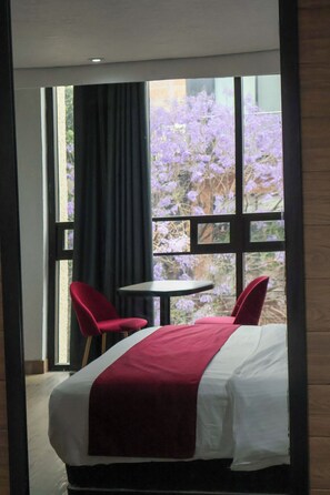 Habitación de hotel con cama, una mesa, dos sillas y ventana con vistas a flores moradas.