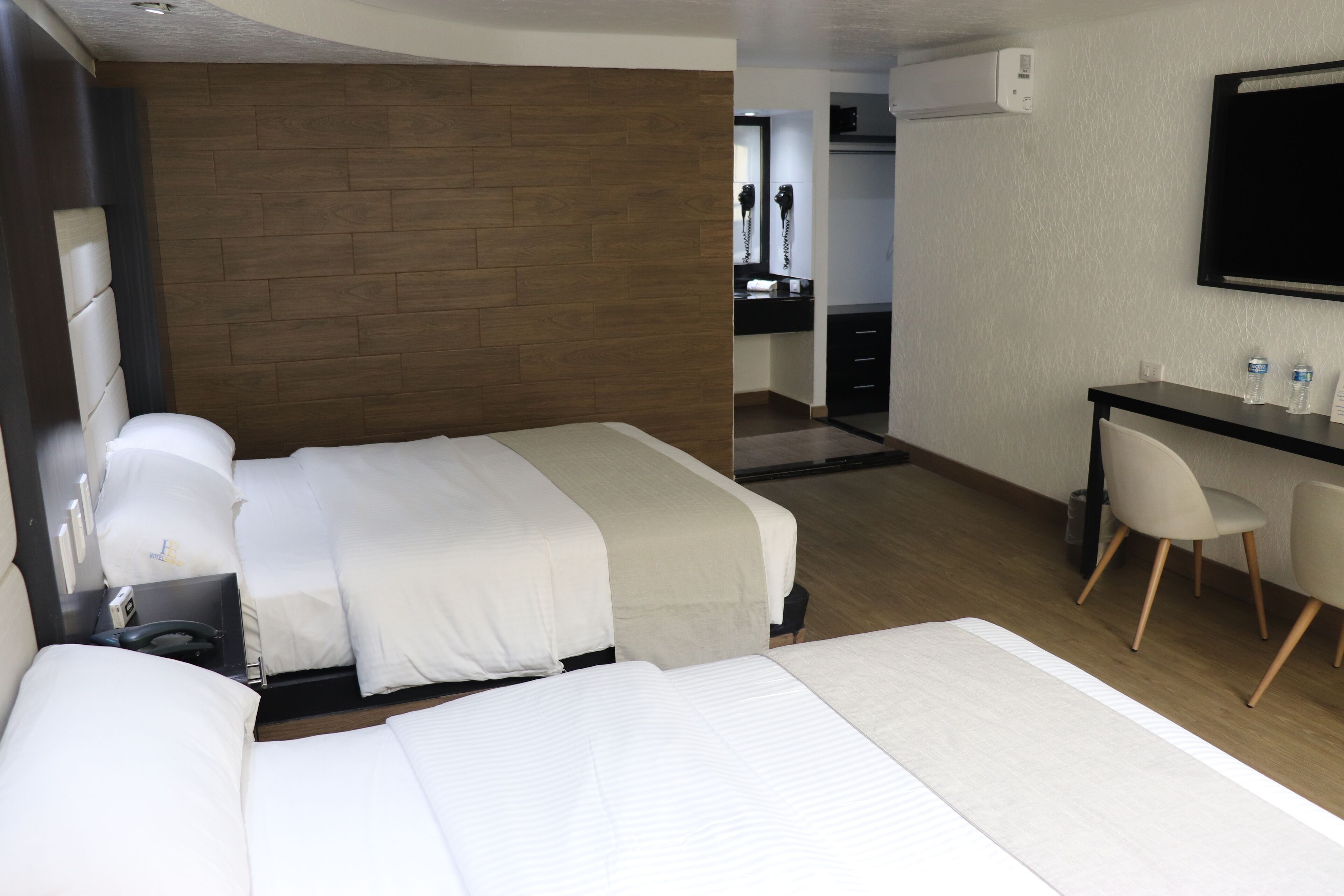 A hotel room with two beds, a desk, and a chair. There is a mirror above the desk and a TV mounted on the wall.