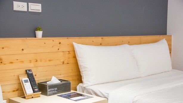 Ein Hotelzimmer mit einem Holzkopfteil, weißer Bettwäsche, einem Nachttisch mit Fernbedienung, einer Taschentuchbox und einer kleinen Topfpflanze an der Wand.