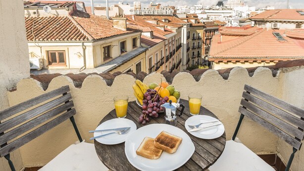 Een balkon met een tafel, ontbijtartikelen, stadszicht en een betegeld dak.