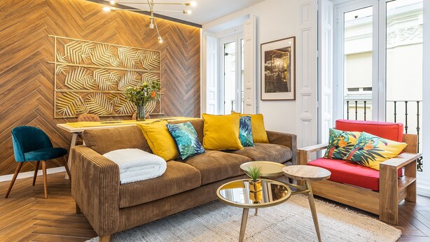 A modern living room with a herringbone wood wall, a sofa with yellow and blue cushions, a red armchair, and a round coffee table. - Waou Sol es MIA - Corta Duración (Madrid)
