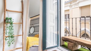 A room with a wooden ladder, a hanging plant, a bed with a yellow blanket, and a view of a building with balconies. - Waou Sol es MIA - Corta Duración (Madrid)