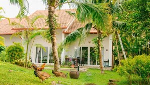 Un giardino tropicale con un edificio bianco dotato di tetto in tegole, circondato da palme e vegetazione lussureggiante.