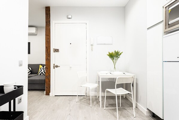 Une petite pièce aux murs blancs, avec une porte, une table blanche et deux chaises, une petite lampe de table blanche et une plante en pot.