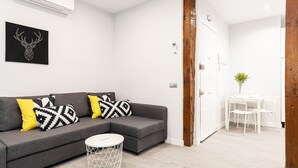 A modern living room with a grey sectional sofa, white and patterned cushions, a small round table, and a white wall-mounted air conditioner.