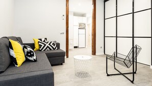 A modern living room with a grey sofa, yellow and black patterned cushions, a round white side table, and a wireframe chair.