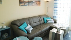 Ein gemütliches Wohnzimmer mit einem braunen Ledersofa, türkisblauen Kissen, einem kleinen runden Tisch und einem Beistelltisch mit dekorativen Gegenständen.