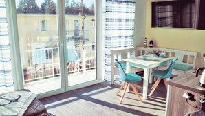 Ein heller Raum mit einer Schiebetür zu einem Balkon, einem weißen Tisch mit blauen Stühlen und einem an der Wand montierten Fernseher.