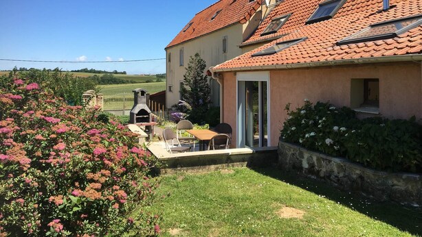 Une maison avec un toit recouvert de tuiles rouges, une terrasse avec une table et des chaises, et un jardin parsemé de fleurs roses.