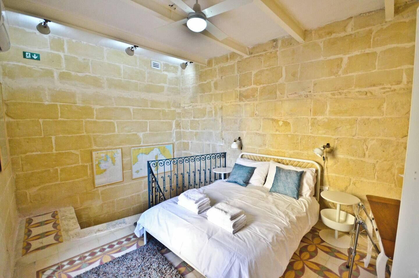 Une chambre avec un mur en pierre, un lit recouvert de draps blancs, une petite table de chevet et une carte accrochée au mur.