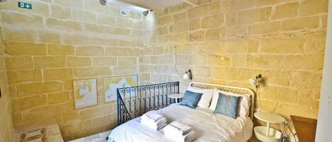 Une chambre avec un mur en pierre, un lit recouvert de draps blancs, une petite table de chevet et une carte accrochée au mur.