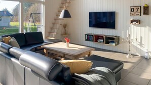 Una sala moderna con un sofá de cuero negro, una mesa de centro de madera y un televisor montado en la pared. Se observan estanterías y una escalera apoyada contra la pared.