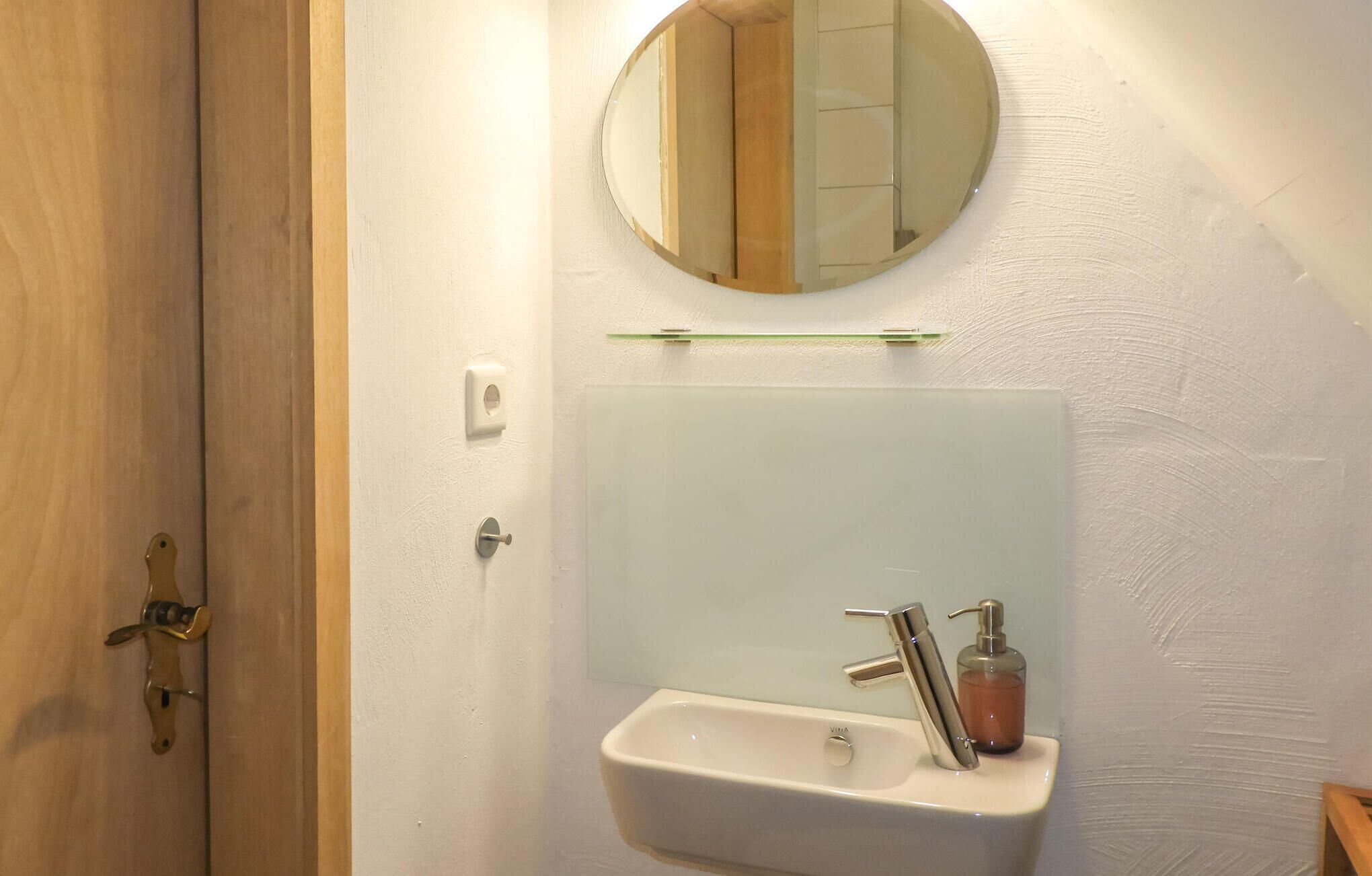 Un bany amb una porta de fusta, un lavabo blanc amb dosificador de sabó, un mirall i un interruptor de llum.
